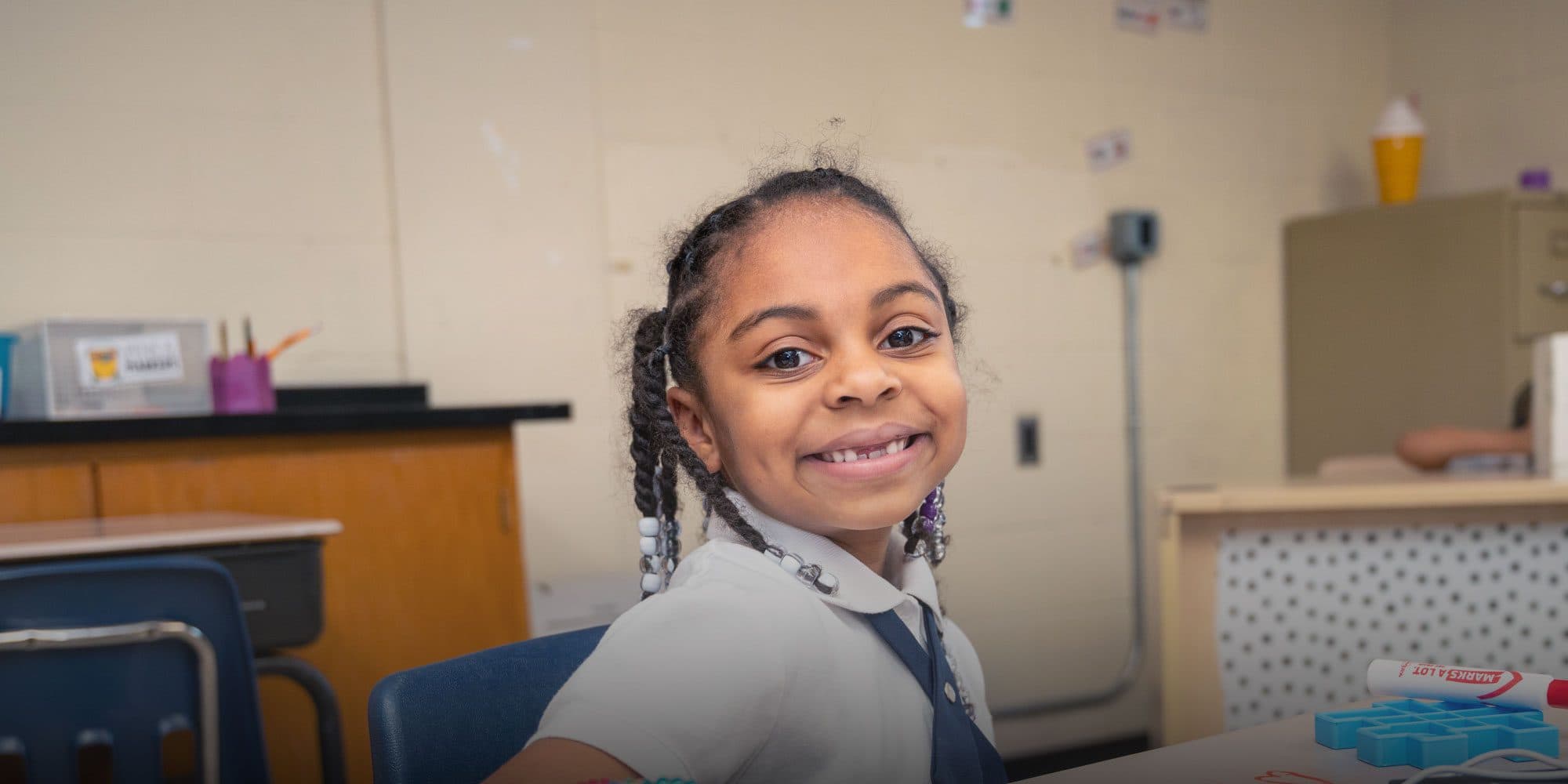 Smiling young student with braided hair seated at a classroom desk.