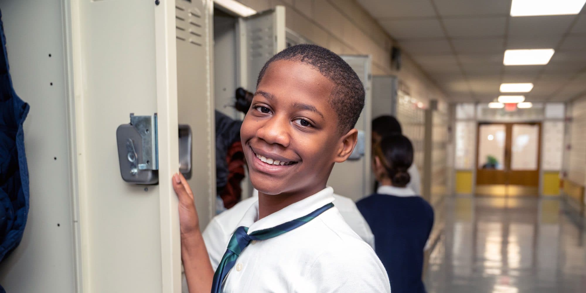 Smiling student in a school uniform and tie standing at an open locker in a school hallway.