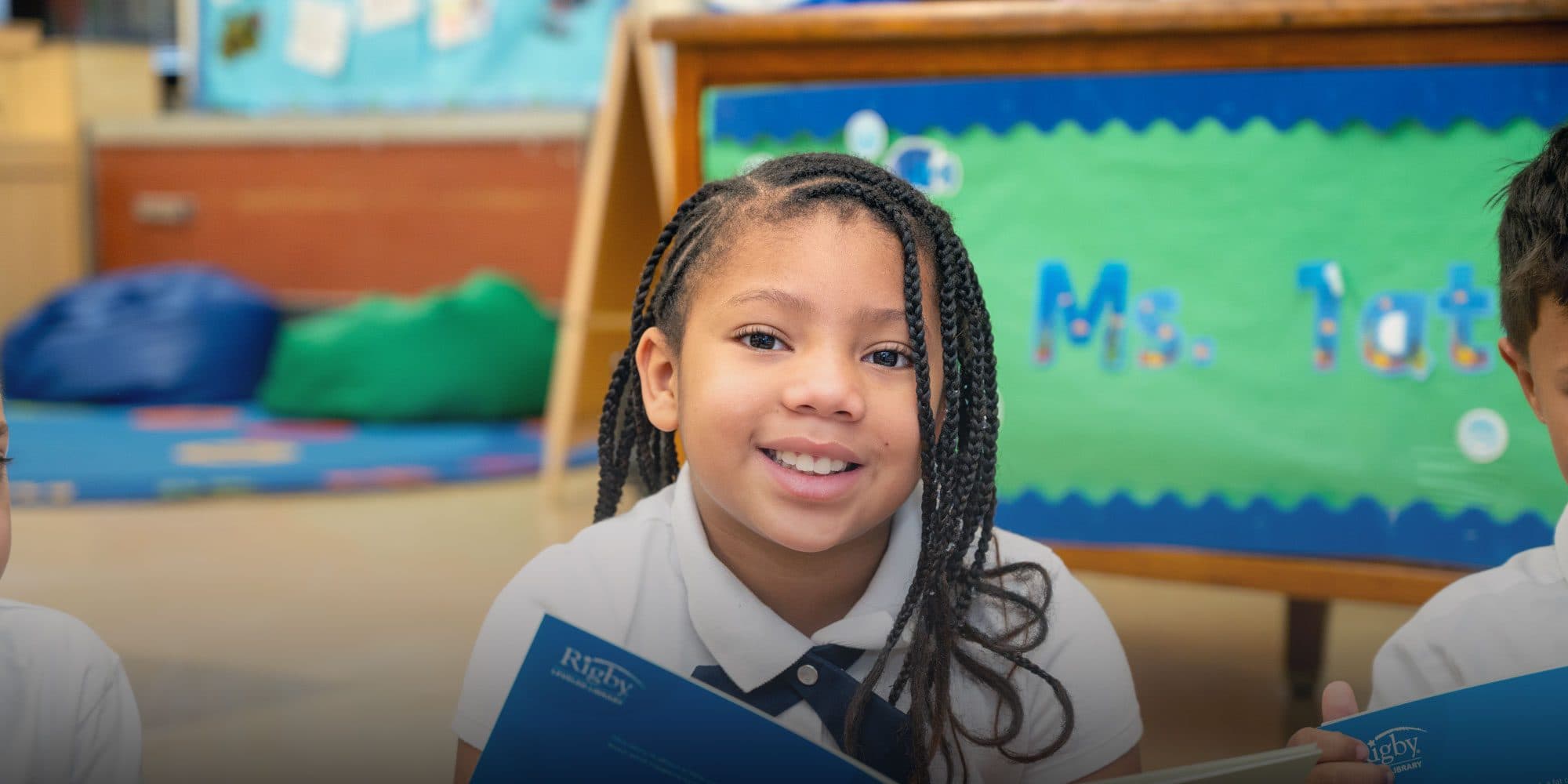 Smiling student with braided hair holding a book in a classroom.