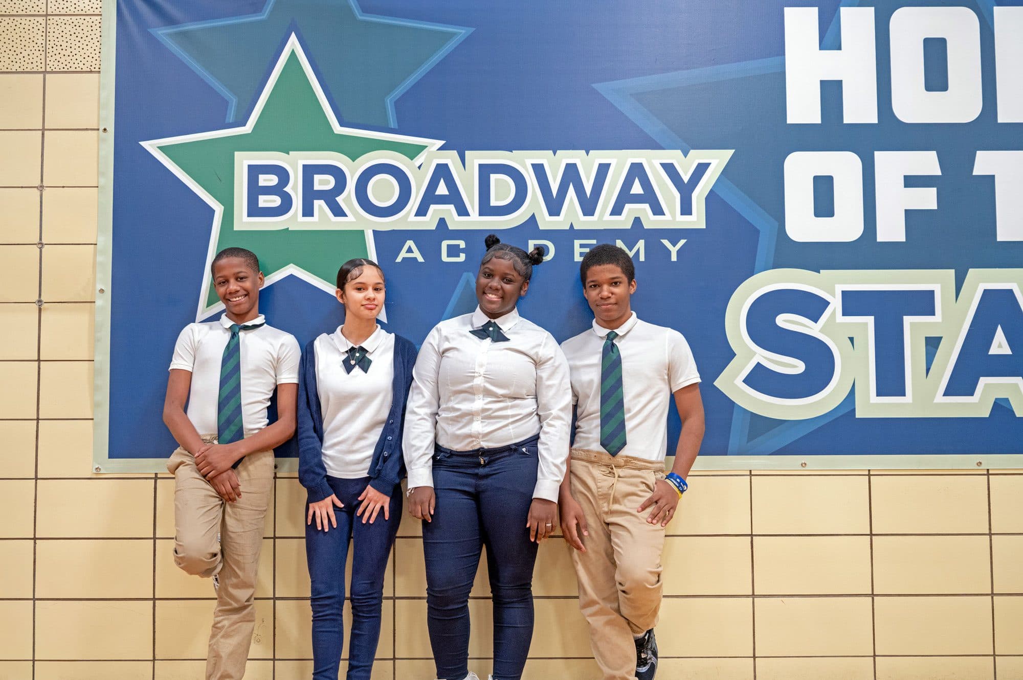 Four students in school uniforms standing in front of a Broadway Academy banner.