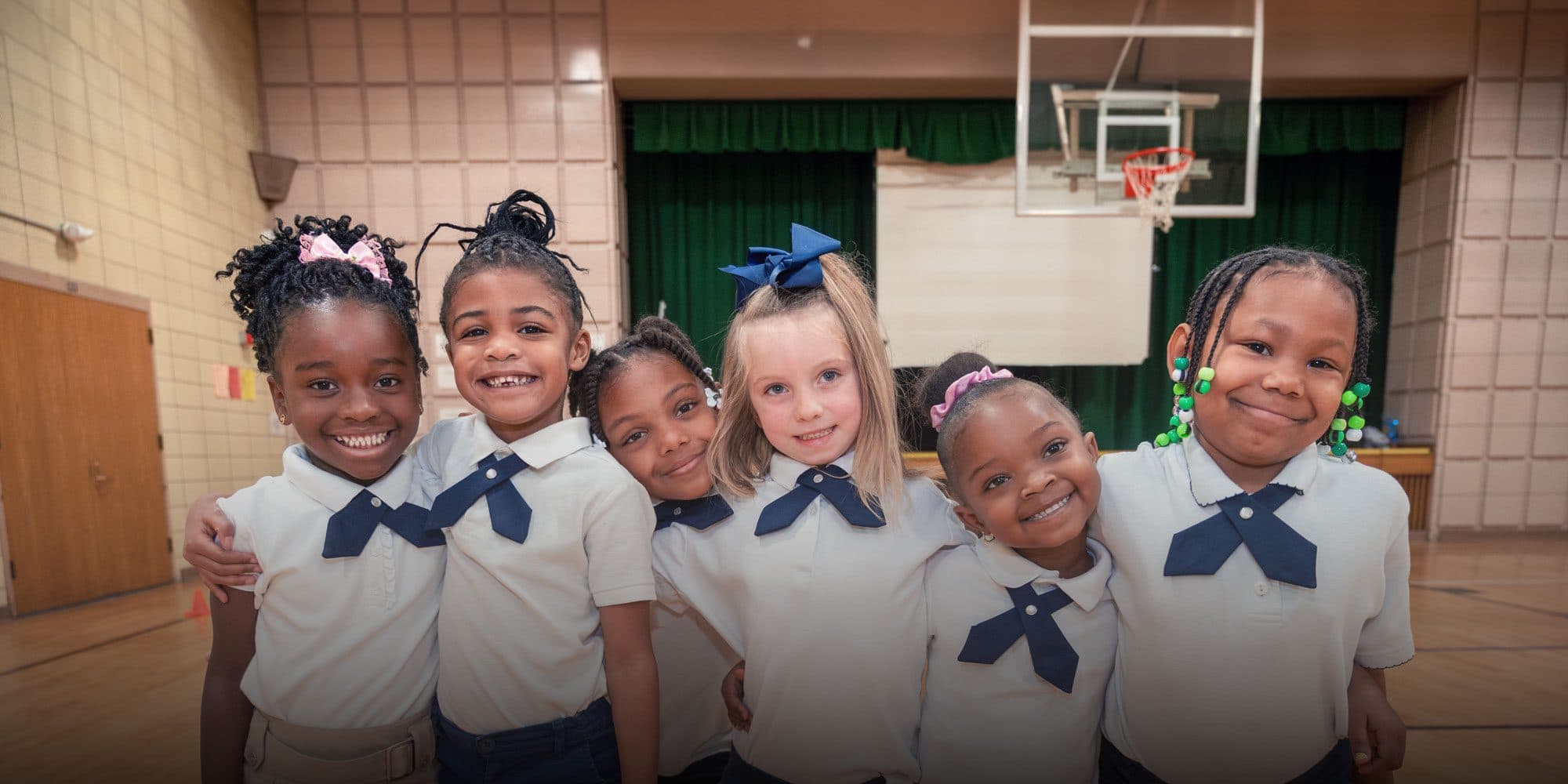 Six smiling students in school uniforms standing arm in arm in the gymnasium.