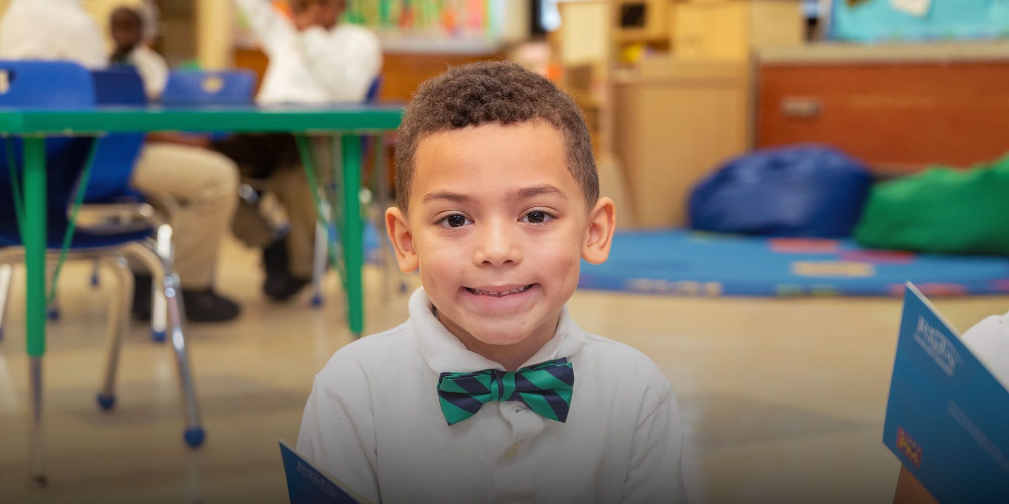 Smiling young student in a green and navy striped bow tie in a classroom.