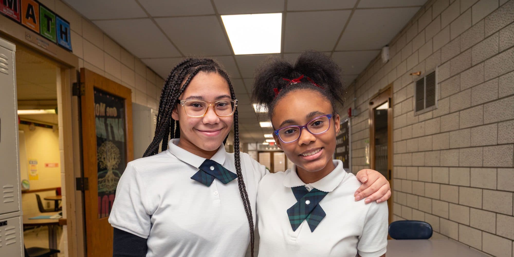 Two smiling students in glasses and school uniforms standing together in a school hallway.