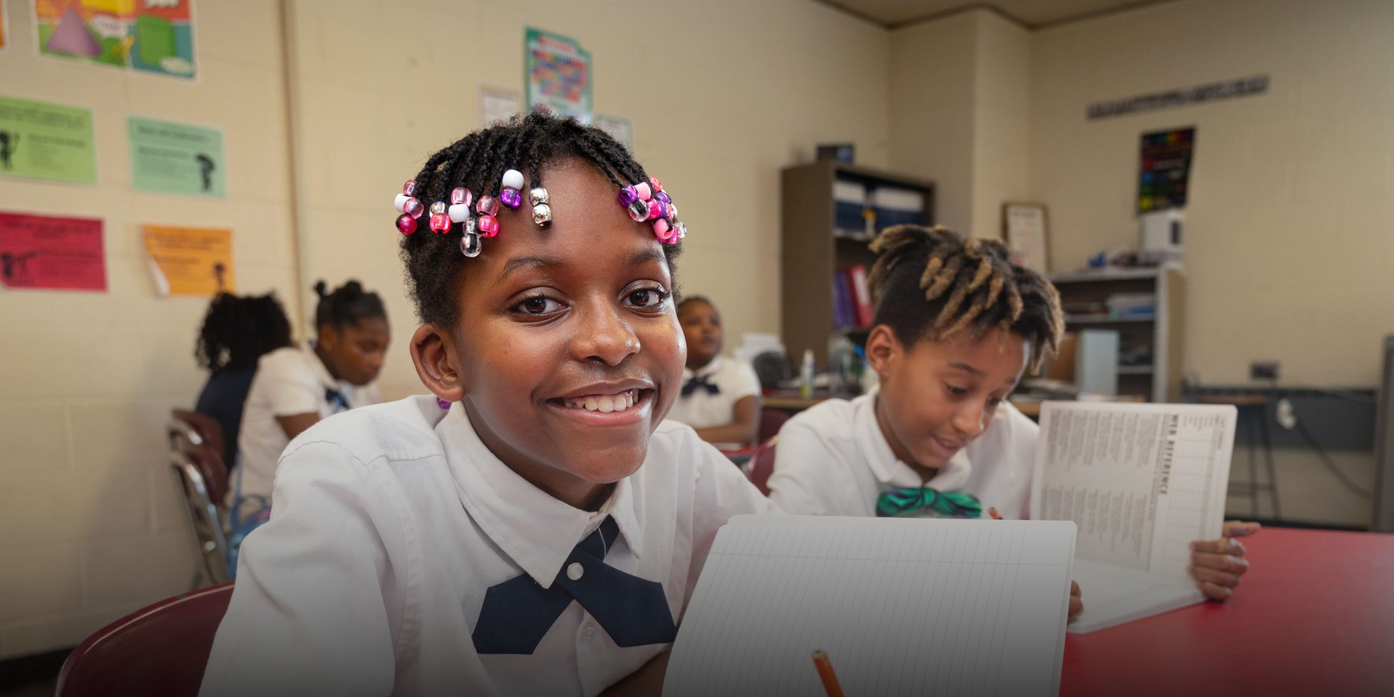 Smiling student with beaded braids working at a desk with a notebook in a classroom.