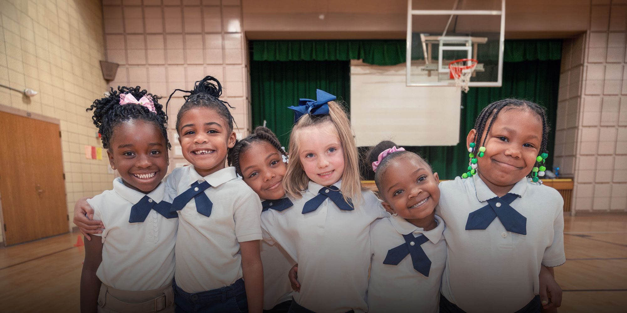 Six smiling students in school uniforms standing arm in arm in the gymnasium.