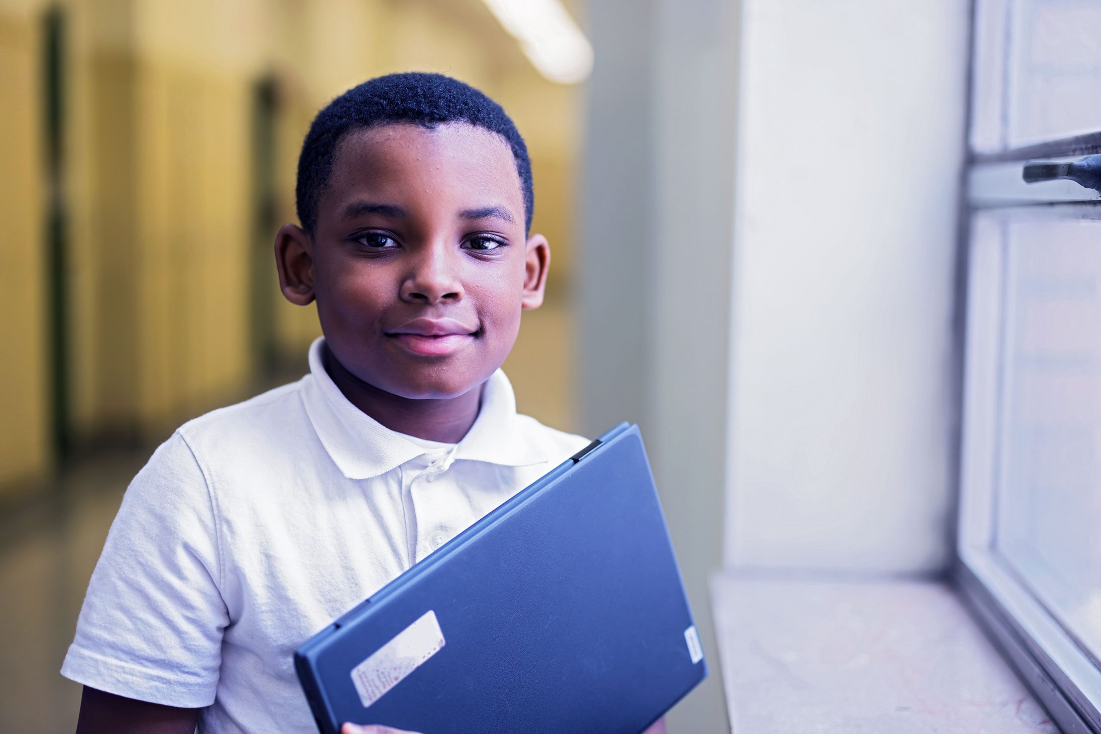 Smiling student holding a laptop in school hallway.